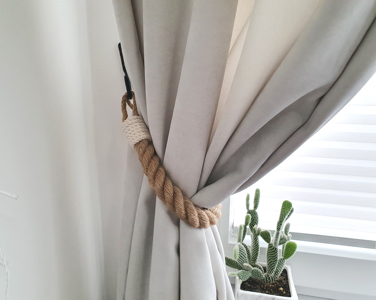 A light gray curtain gathered back with a natural jute rope tie and a black hook, shown in a bright room next to a window with blinds and a small potted cactus.