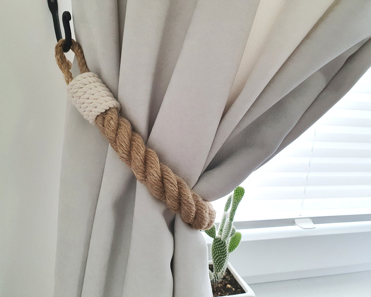 A rope curtain tieback with a natural jute twisted handle and a cream knotted wrap, hanging from a black hook on a light gray curtain next to a window with blinds and a small cactus plant.