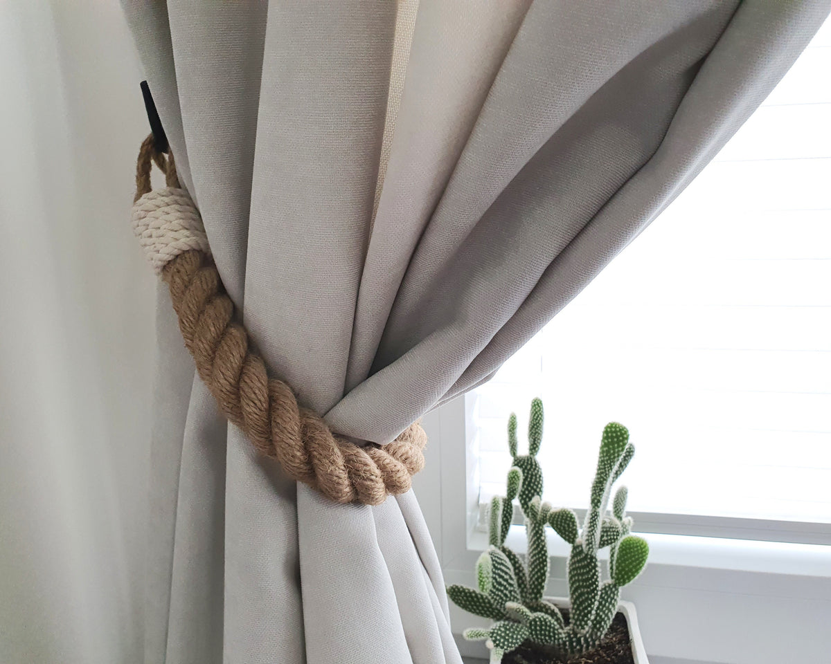 A beige curtain panel tied back with a thick twisted rope tieback, shown next to a bright window with a small potted cactus in the foreground.