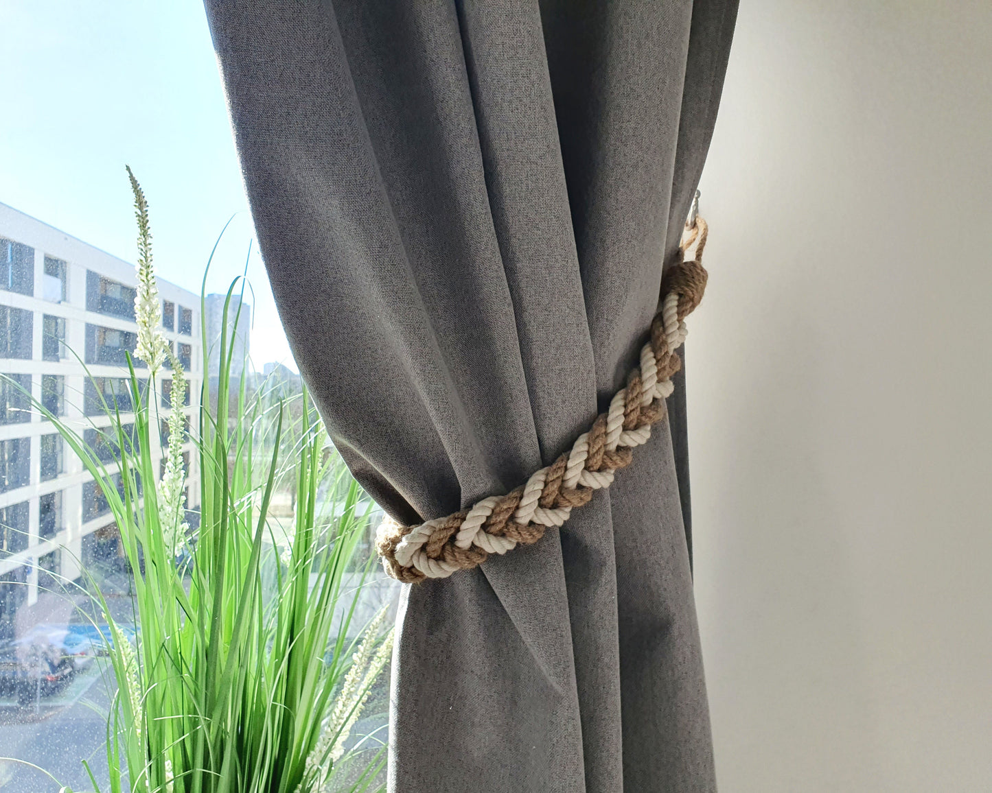 Gray curtain with a braided rope tieback in front of a window with a view of buildings and greenery.