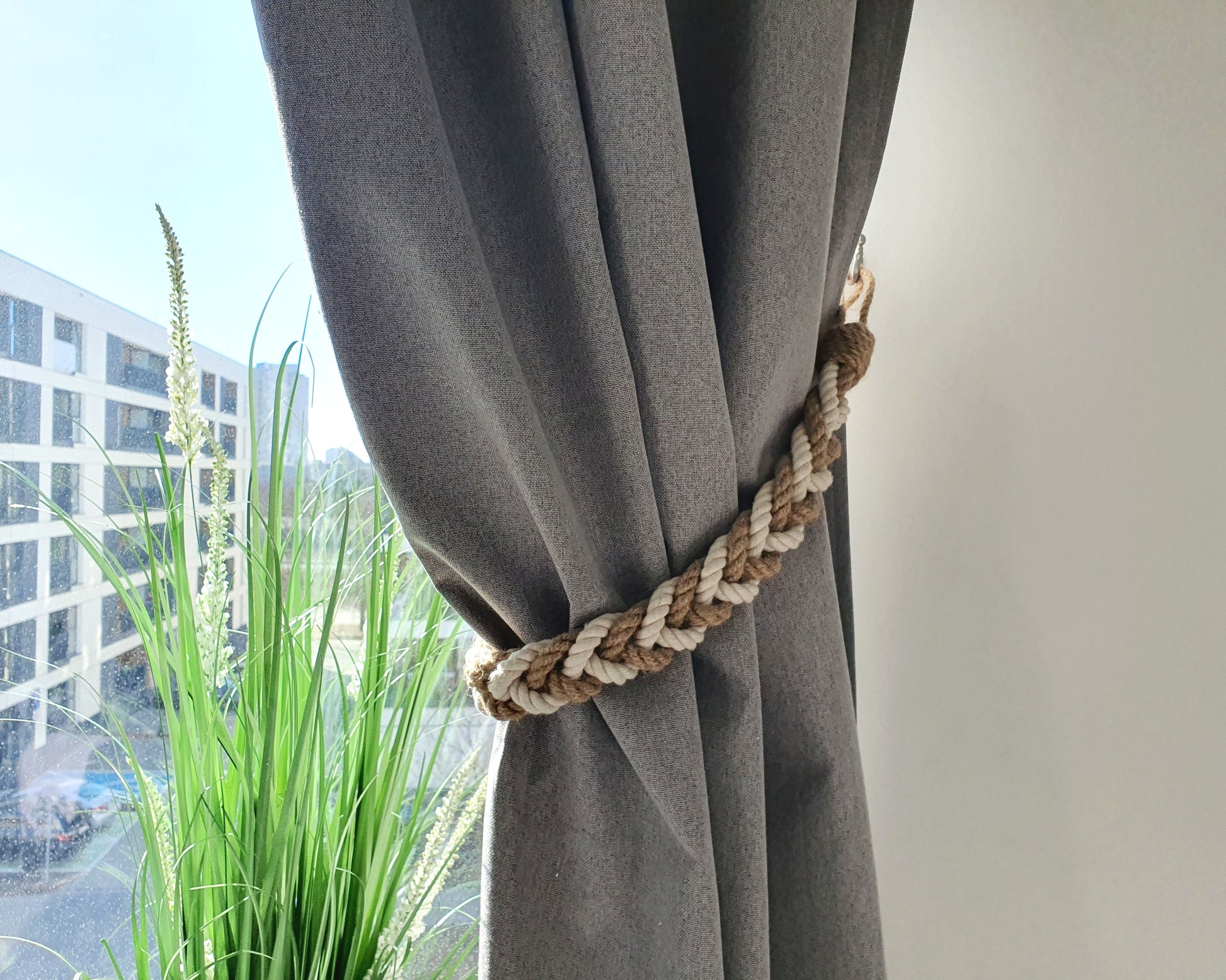 Gray curtain with a braided rope tieback in front of a window with a view of buildings and greenery.