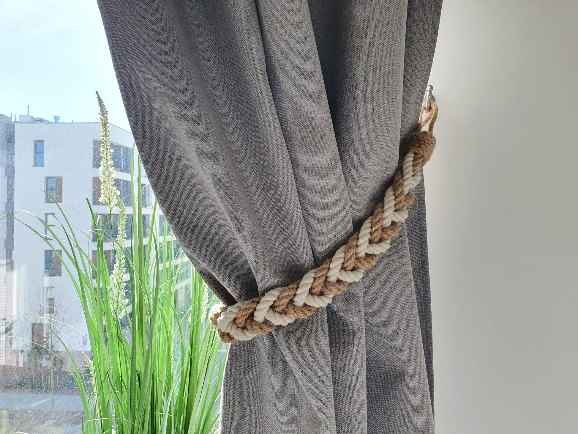 Gray curtain with a decorative rope tieback in front of a window with greenery outside.