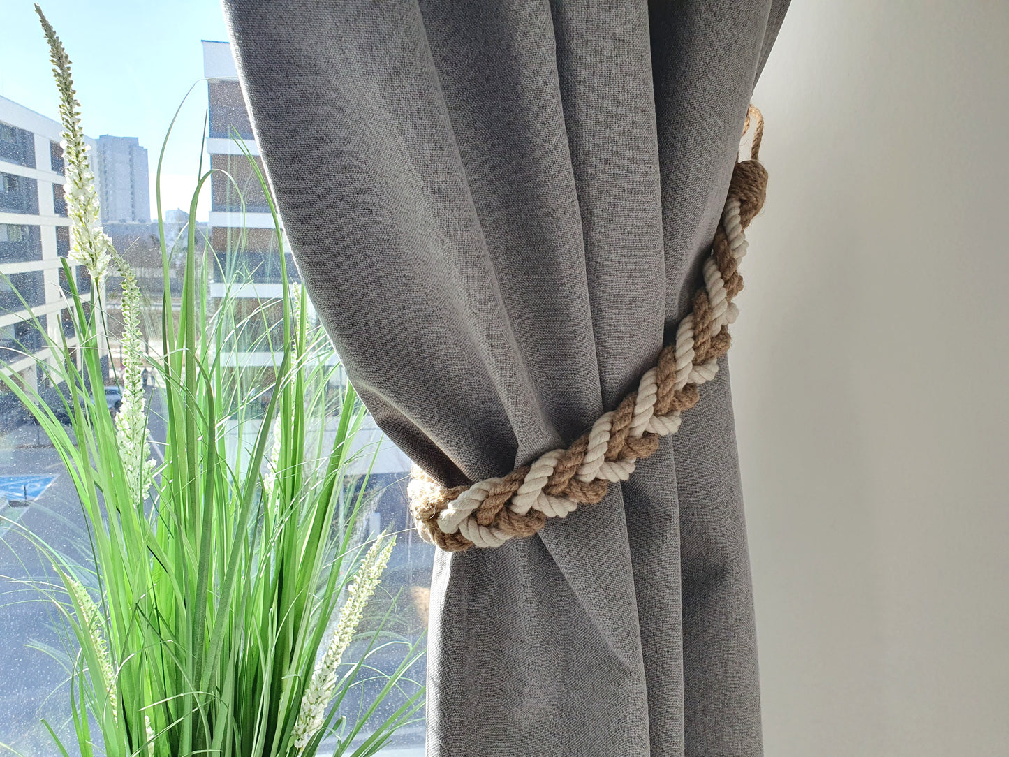 Gray curtain with a decorative rope tieback in front of a window with green plants.