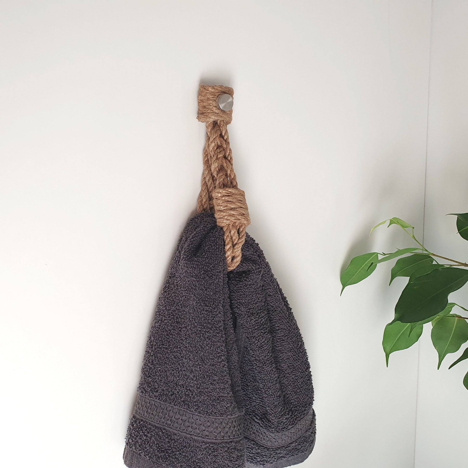 A dark gray textured towel hanging from a natural jute rope wall hook on a white wall, with green plant leaves visible on the right side.