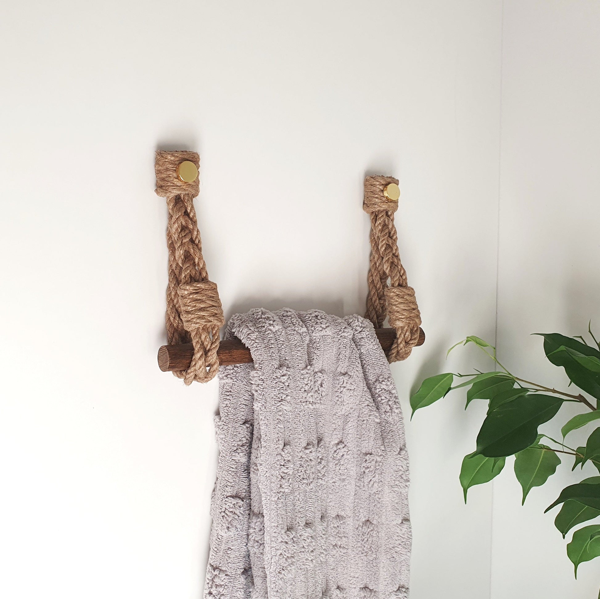 A light gray textured throw blanket draped over a rustic wooden towel rack made from two wrapped rope handles, mounted on a plain white wall with a green plant visible in the corner.