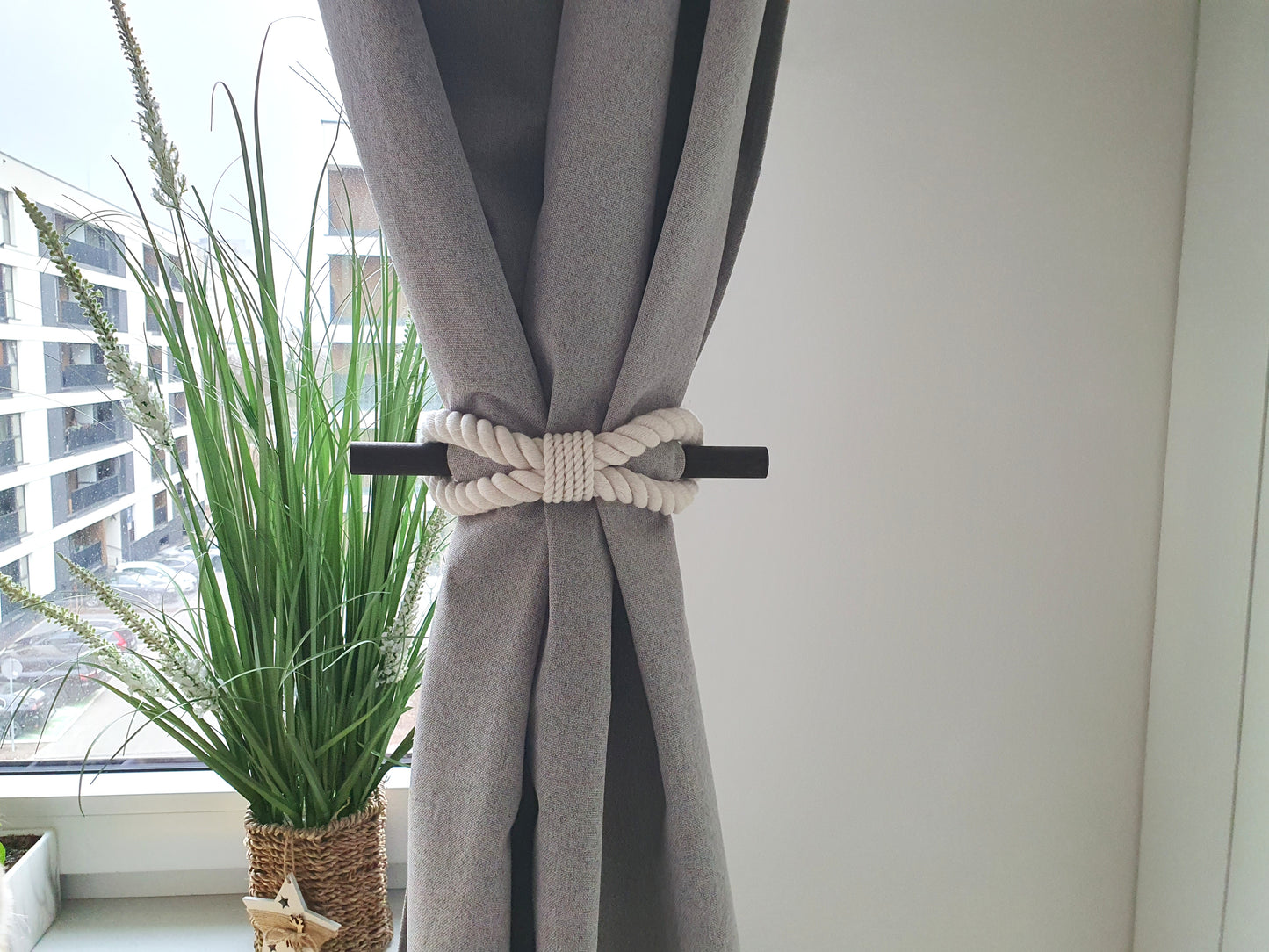 Grey curtain with a decorative white and black tieback in a room with a plant and window view.