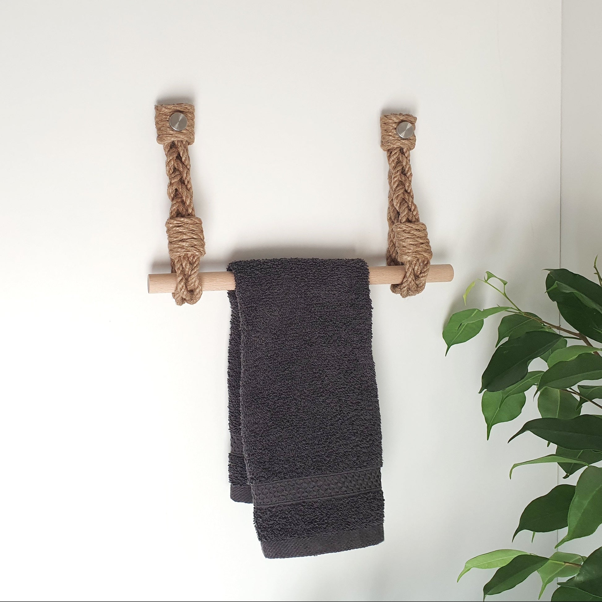 A dark charcoal gray towel hangs from a rustic rope-and-dowel towel rack mounted on a white wall, with a gray curtain on the left and a green houseplant on the right.