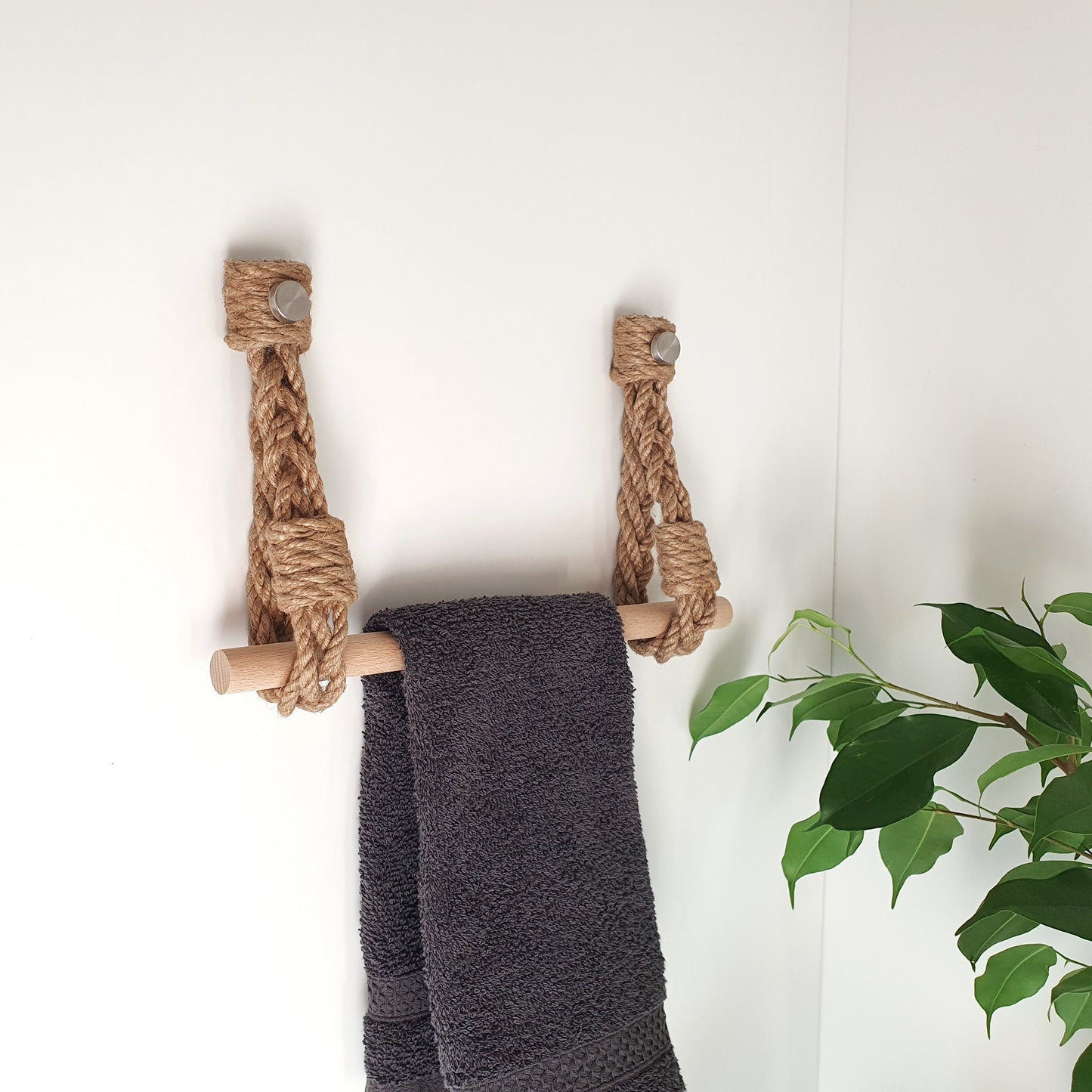 A rope ladder towel rack holder made of natural jute and light wood, holding a dark gray towel, mounted on a white wall with a green plant visible on the right.
