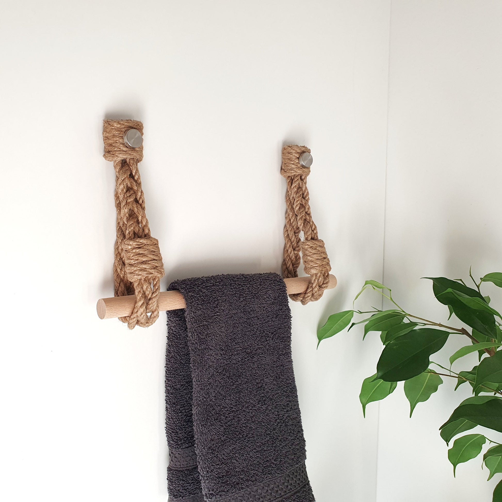 A rope ladder towel rack holder made of natural jute and light wood, holding a dark gray towel, mounted on a white wall with a green plant visible on the right.