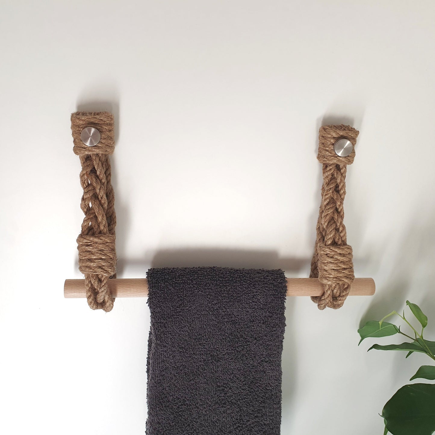 A rustic rope towel bar with a natural wood rod and silver metal wall anchors, holding a dark gray towel against a plain white wall.