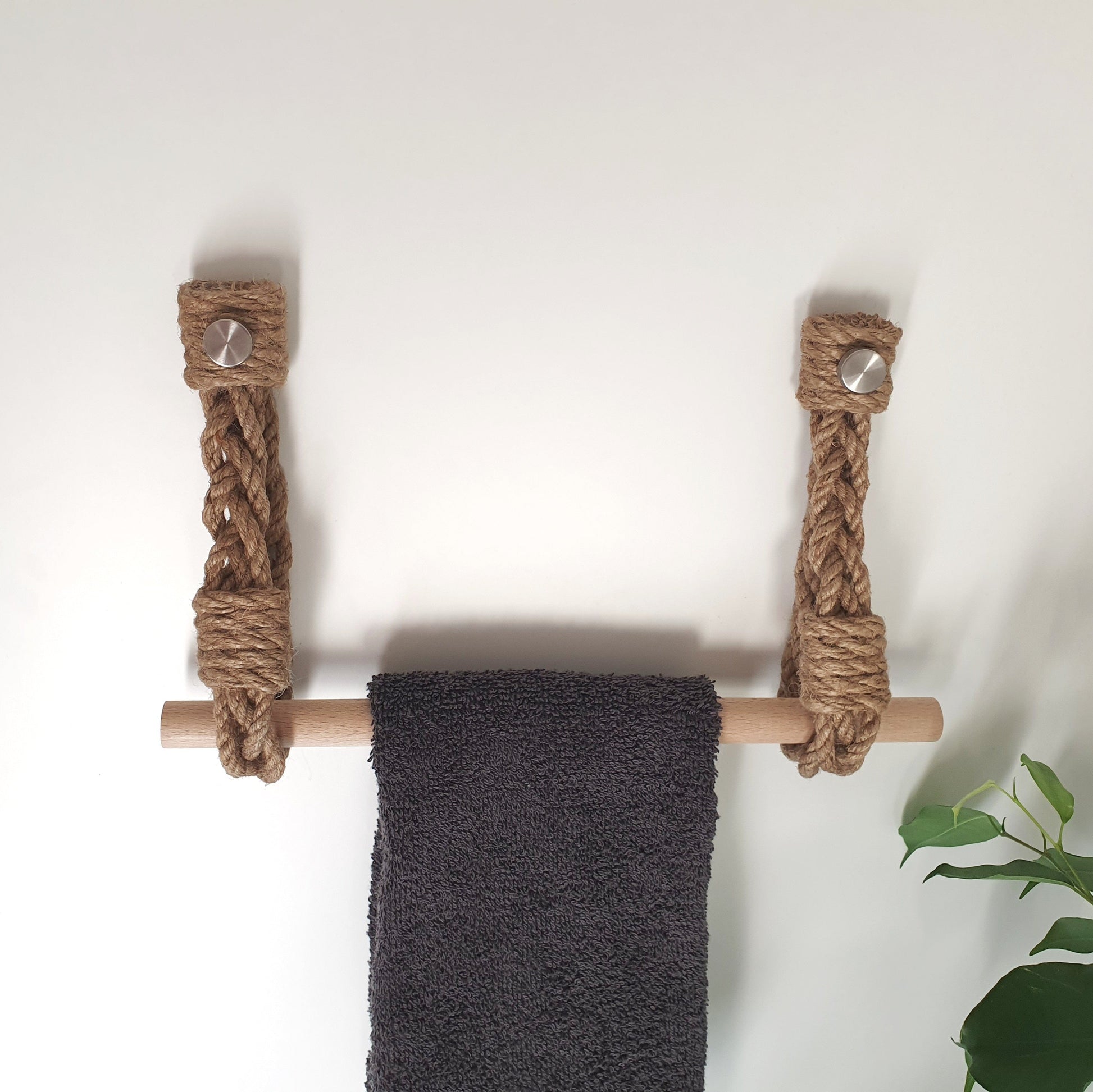 A rustic rope towel bar with a natural wood rod and silver metal wall anchors, holding a dark gray towel against a plain white wall.