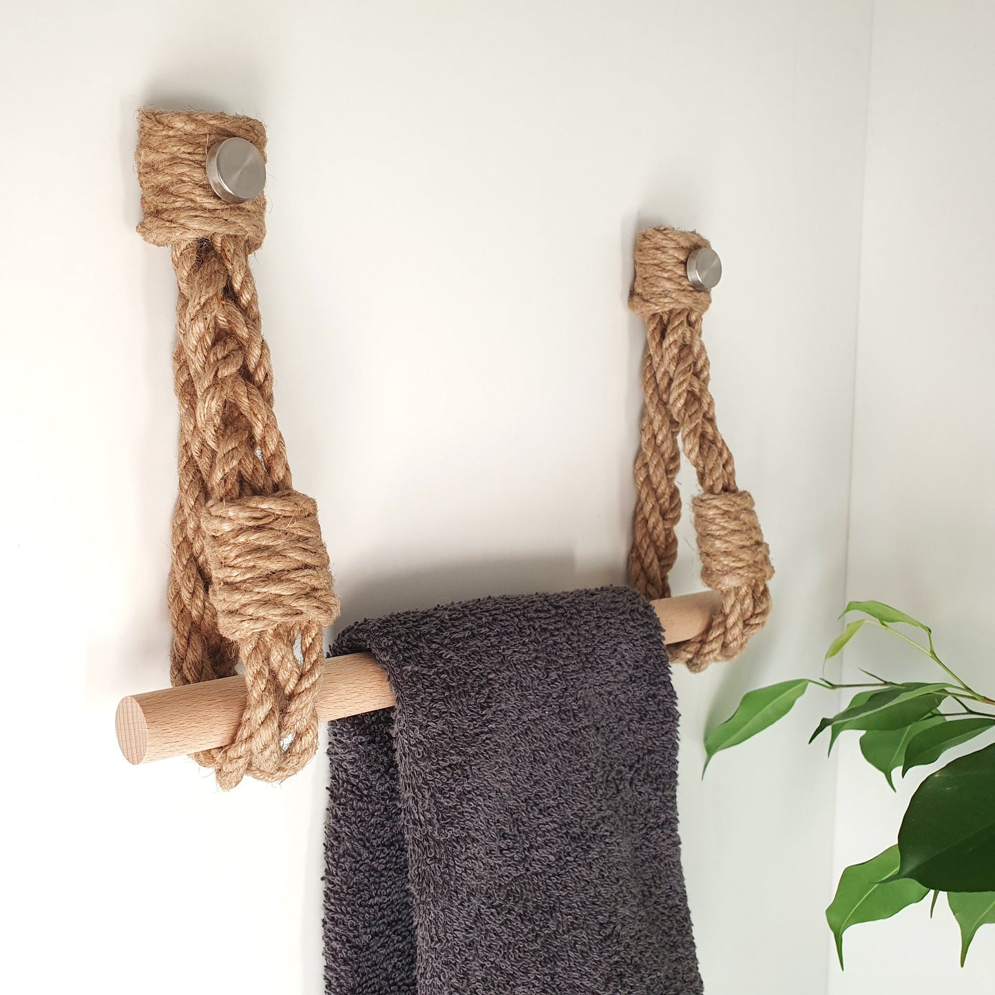 A rope towel rack with wooden bars and metal wall hooks, shown hanging on a white wall with a dark gray towel draped over it and a small green plant leaf in the corner.