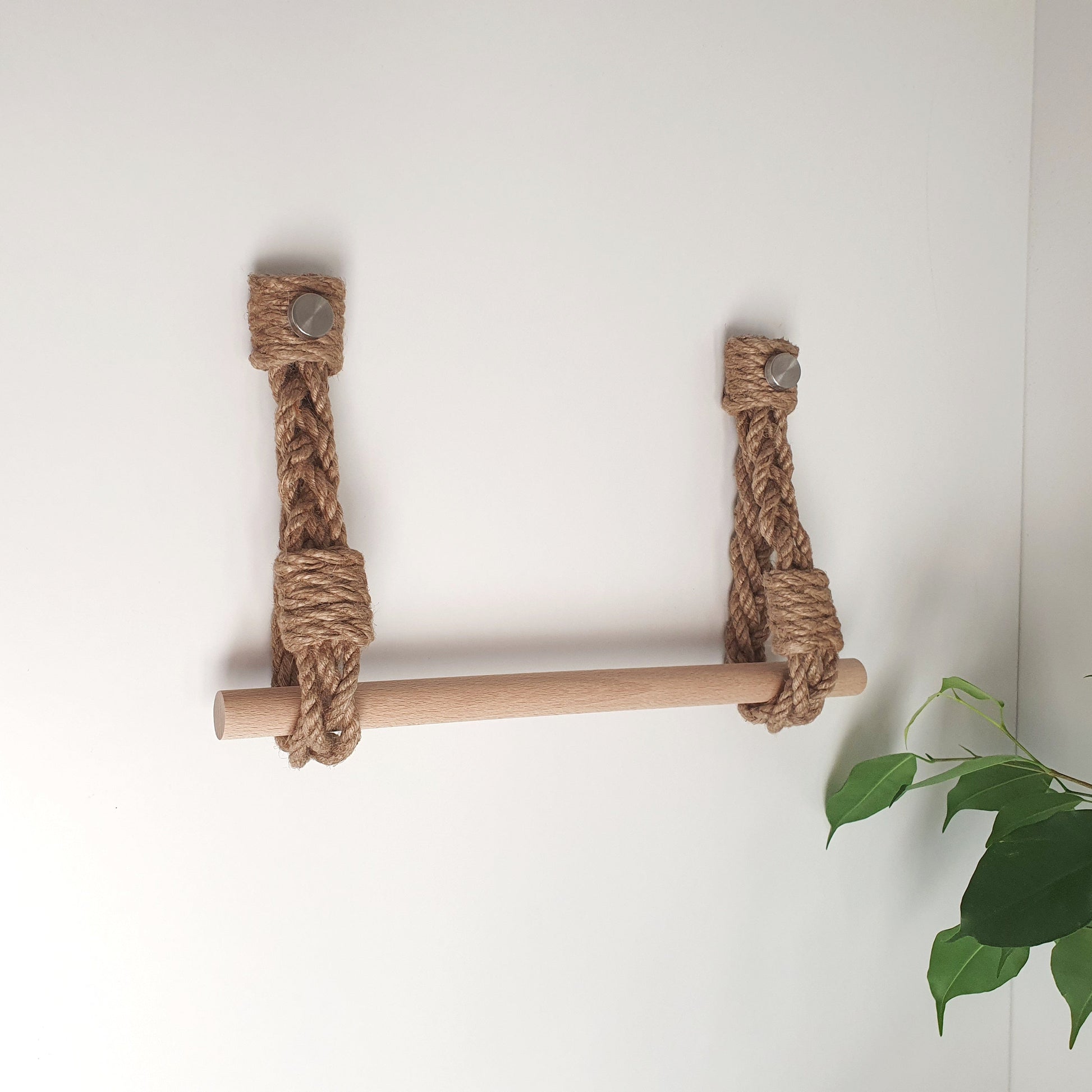 A rustic wall-mounted wooden ladder shelf with thick natural jute rope wrapping around the side supports, shown against a plain white wall with a green plant leaf visible in the lower right corner.