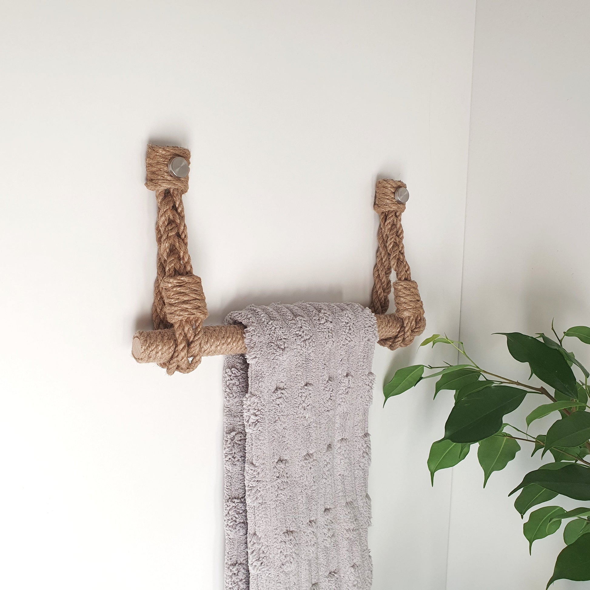 A natural jute rope towel rack hanging on a white wall, holding a textured light gray towel, with a green plant leaf visible in the lower right corner.