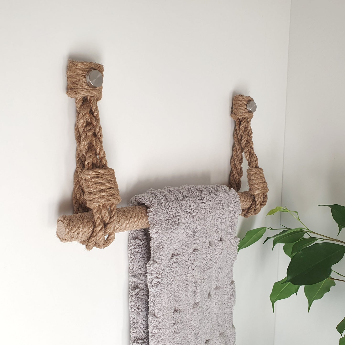 A rustic rope towel rack mounted on a white wall, holding a textured light gray towel. A green houseplant is visible in the lower right corner.