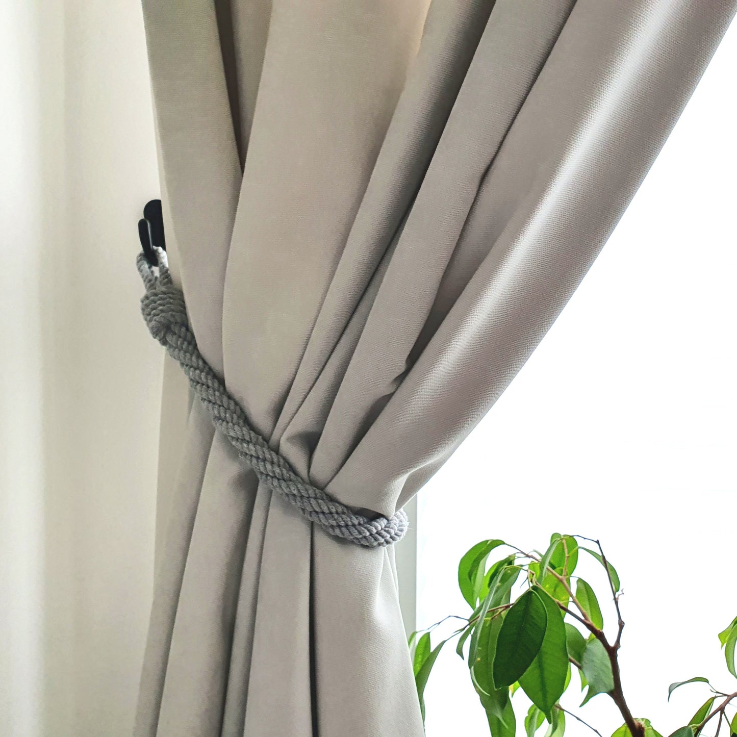 A close-up of a light beige curtain tied back with a gray braided rope tieback, with a bright window background and green plant leaves visible in the lower right corner.