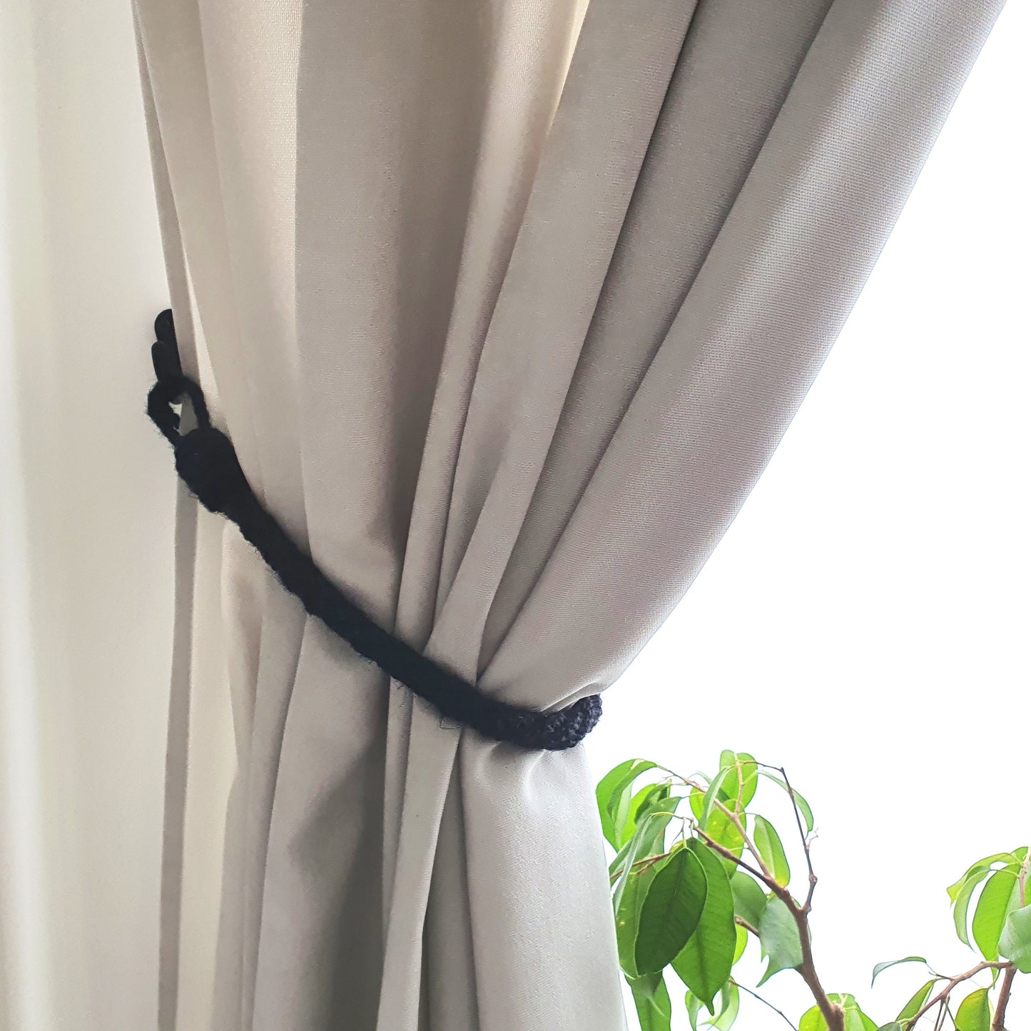 A close-up of a light beige curtain tied back with a black rope curtain buster, with a bright window background and green plant leaves visible in the foreground.