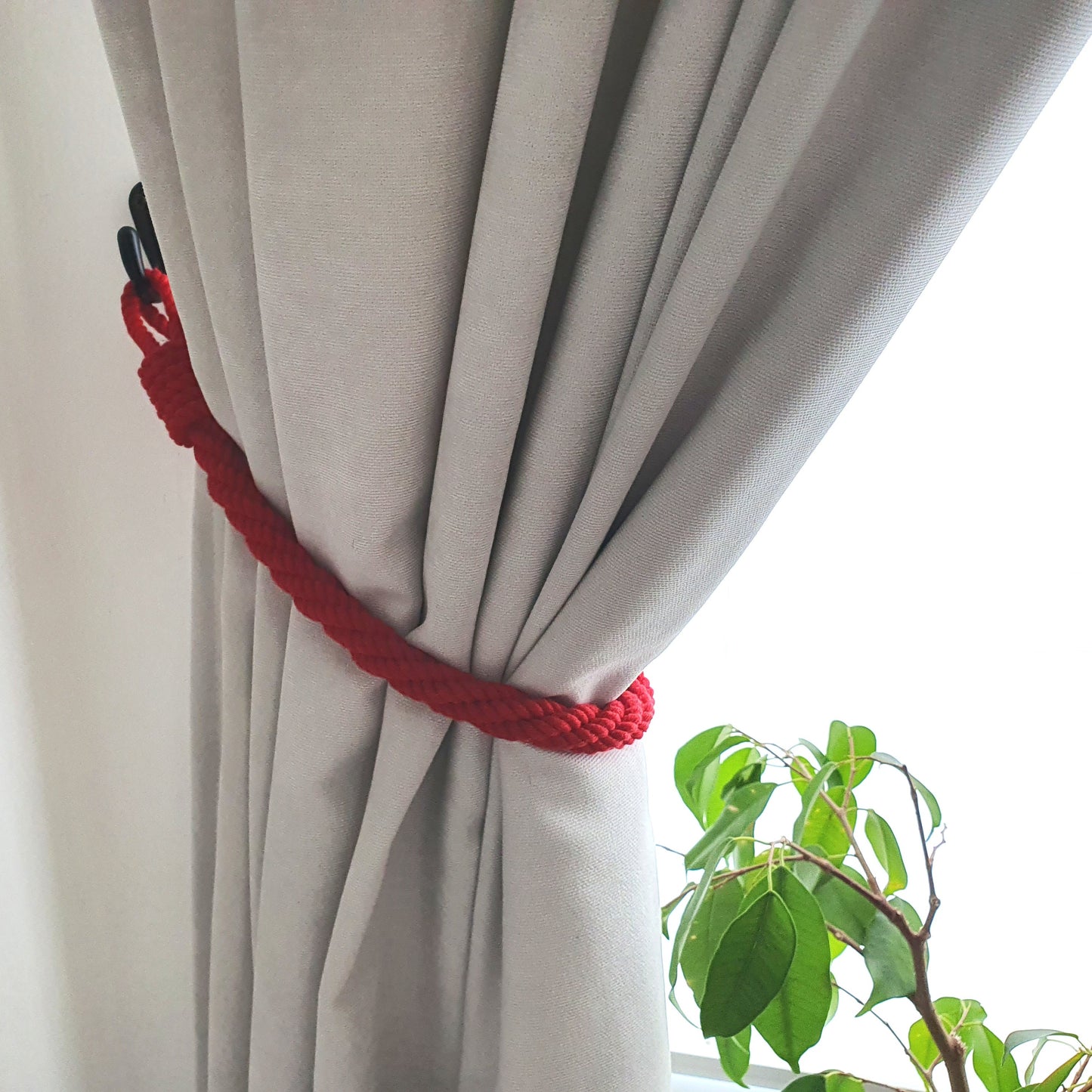 A light gray curtain tied back with a bright red braided rope tieback, shown next to a window with bright daylight and a green plant in the foreground.