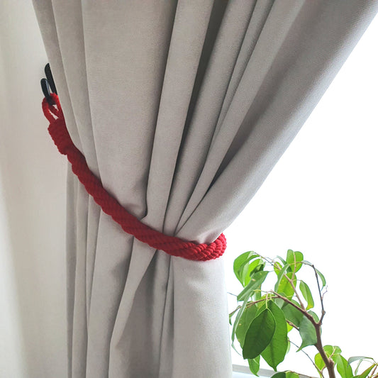 A light gray curtain tied back with a bright red braided rope tieback, shown next to a window with bright daylight and a green plant in the foreground.