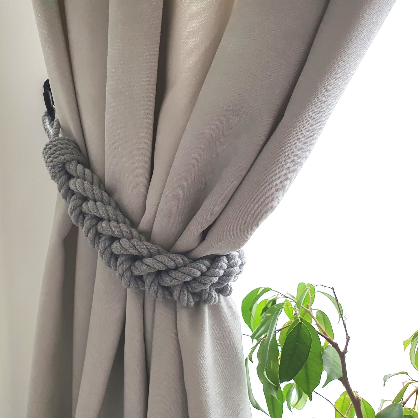 A gray braided rope curtain tieback holding back a light beige curtain, with a bright window background and green plant leaves visible in the lower right corner.