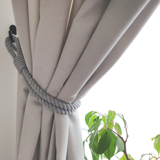 A light gray rope curtain tieback holding back a beige curtain panel, with a bright window background and a small green plant visible in the lower right corner.